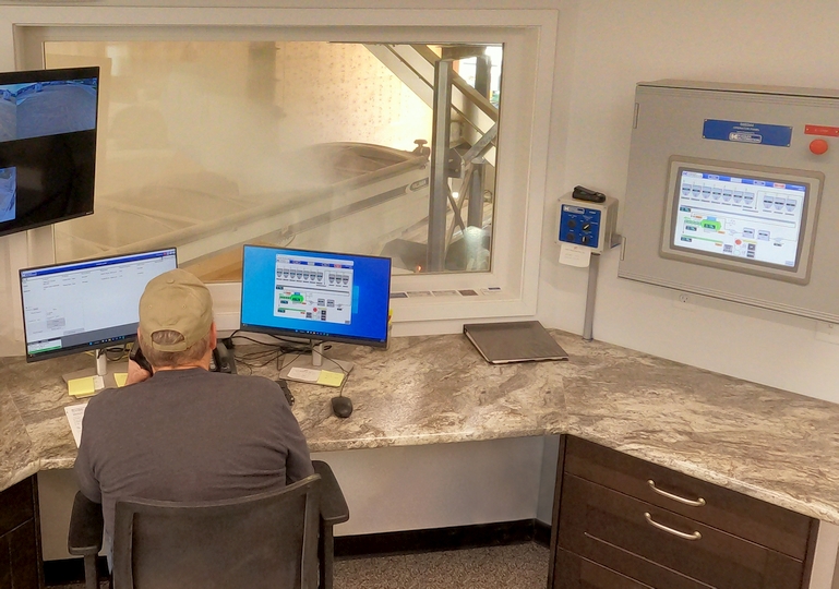 Blend plant operator at the controls in a Fertilizer building control room with windows while loading dry plant food blends